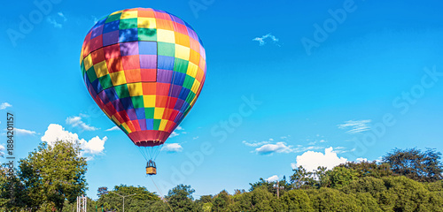 Vibrant multicolored checkered hot air balloon floating gracefully in a clear blue sky over lush greenery.