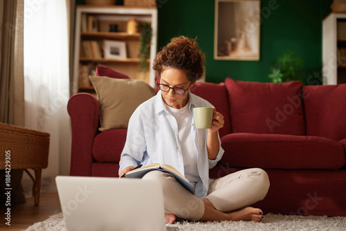 Photography A woman sits cross-legged on the floor with a cup of coffee in hand, absorbed in a book while using a laptop in a warm, inviting living room, surrounded by soft furnishings and greenery