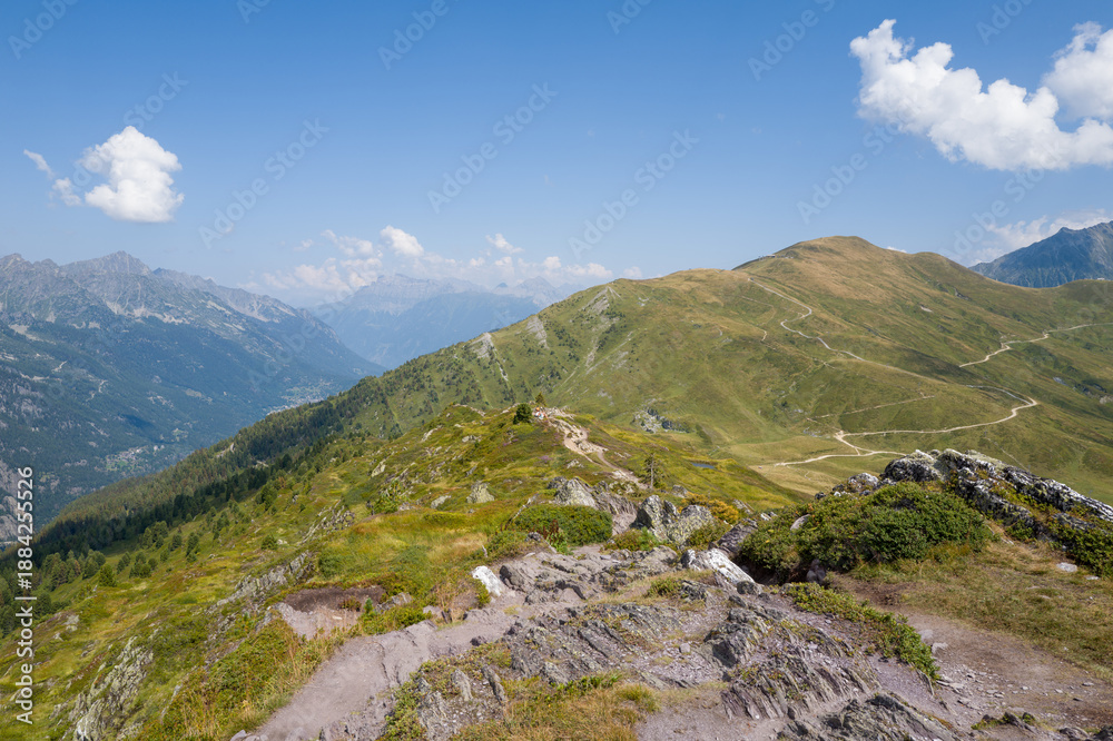 Obraz premium Rocky mountain ridge overlooks rolling green slopes crisscrossed by winding paths under a clear blue sky in the Aiguillettes des Posettes region.