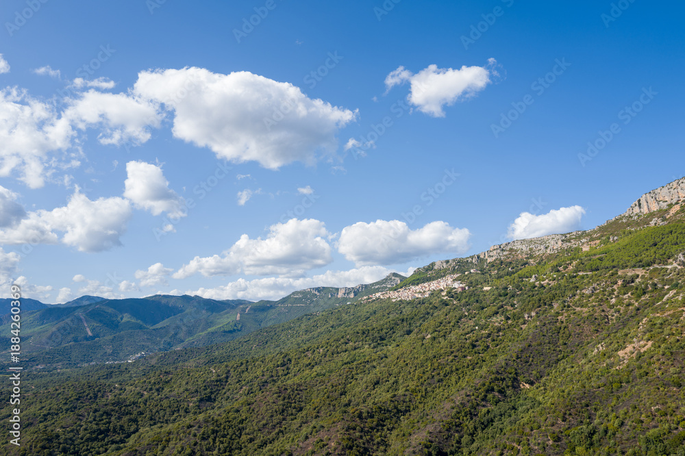 Fototapeta premium Vast rolling hills blanketed in lush greenery stretch beneath a bright blue sky dotted with scattered white clouds near Baunei, Sardinia. Sunlight enhances the textures of the rugged Mediterranean