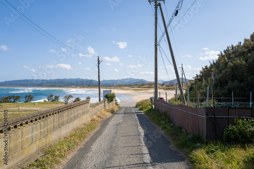 A quiet paved lane bordered by fences and utility poles descends toward the sandy shore of Shotenkyo Beach in Kumihamacho, with blue waves and distant mountains under a clear sky.