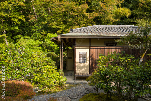 A classic wooden tea house with tiled roof stands surrounded by vibrant green foliage and manicured shrubs in the tranquil gardens of Nanzen-ji, Kyoto, illuminated by gentle afternoon sunlight.