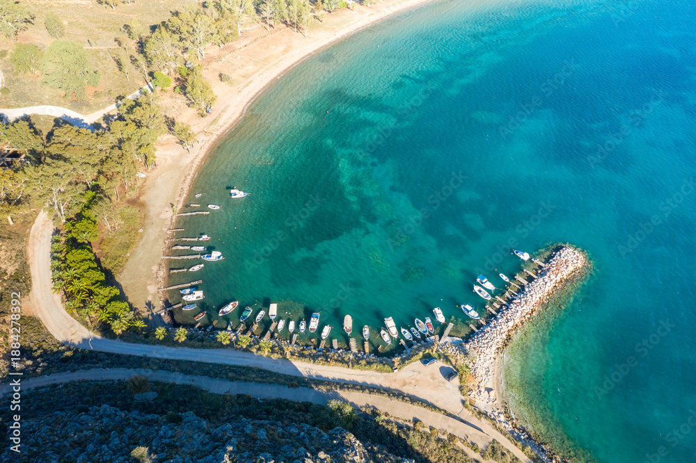 Fototapeta premium Aerial view of a curved bay with a small marina and boats docked along a rocky breakwater at Karathona Beach near Nafplio, Greece. The clear turquoise water contrasts with the sandy shoreline and lush