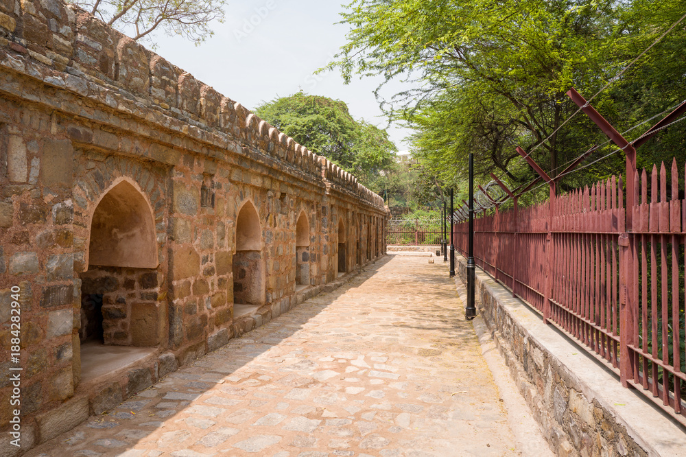 Obraz premium Sunlit stone arcade with arched niches lines a cobblestone walkway in Mehrauli Archaeological Park, Delhi. The historic wall contrasts with iron fencing and dense green trees under a clear sky.