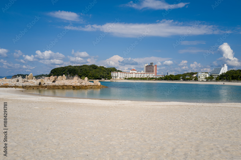Obraz premium A stone jetty extends into calm blue water beside a wide stretch of white sand, with modern hotels and lush green trees under a bright sky in Shirarahama, Japan.