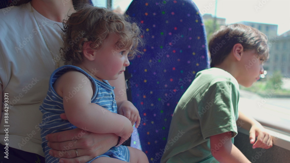 Obraz premium Baby sitting upright in mother’s arms looking out on bus while older child leans against window, both watching passing city scenery
