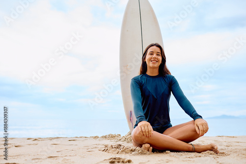 Canvas Print Young woman smiling, wearing a rashguard, sitting crossed-legged on the sand wit