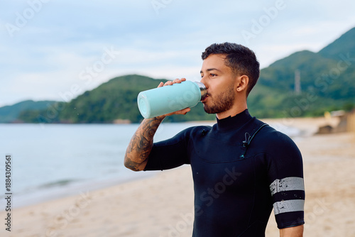 Slika na platnu Surfer man in wetsuit hydrating on a sandy beach, drinking water from a light bl
