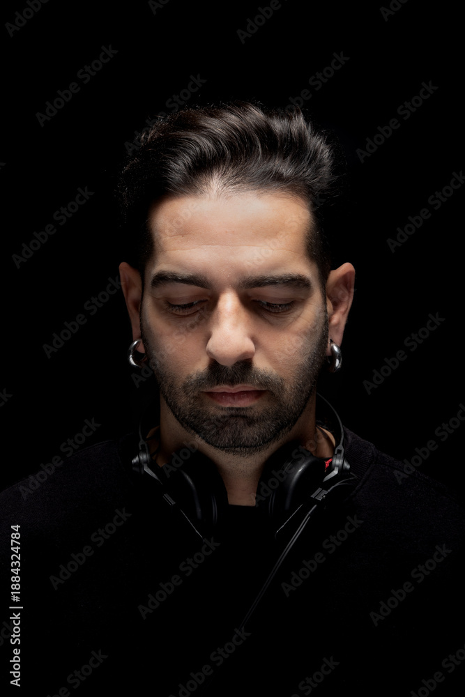 Obraz premium A DJ stands with eyes closed, wearing headphones. The background is dark. He appears to be concentrating on the music as his head is slightly bowed. This moment captures a deep connection to sound.