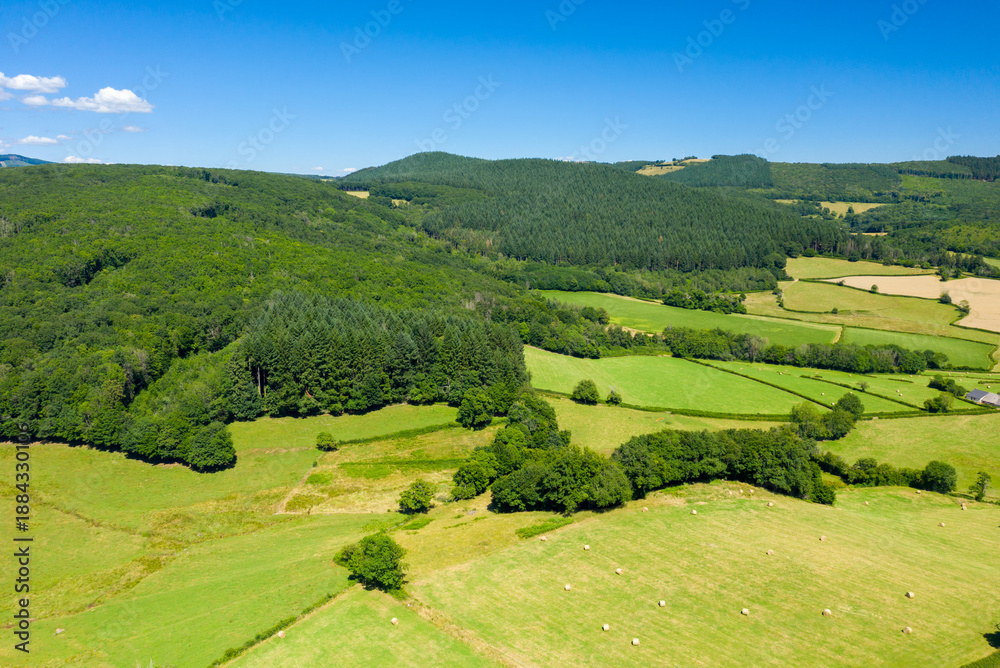 Obraz premium Aerial view of vibrant green hills and dense forests interspersed with geometric fields and scattered hay bales in the Morvan countryside on a clear summer day.