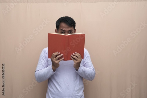 Southeast Asian man wearing a casual long-sleeve t-shirt holding red book covering his face, suggesting privacy, shyness, study focus, or anonymous reader concept for advertising.
