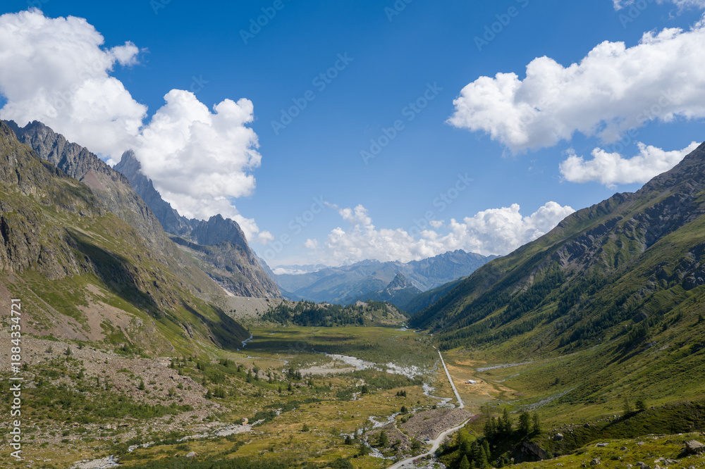 Fototapeta premium Expansive view of a green alpine valley flanked by steep rocky mountains under a bright blue sky with scattered white clouds near Col de la Seigne in the Italian Alps. Sunlight highlights the rugged