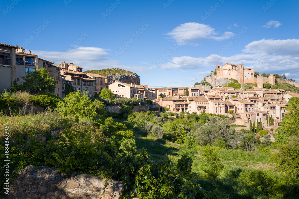 Fototapeta premium Traditional stone houses and a fortified castle rise above lush greenery in the medieval village of Alquezar, Spain. Bright sunlight and scattered clouds create a vibrant, inviting atmosphere across