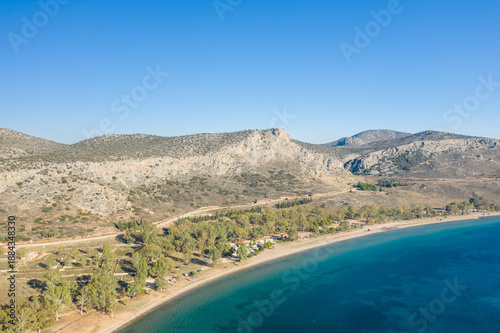 Aerial perspective of Karathona Beach near Nafplio, Greece, showing a quiet sandy shoreline bordered by clear blue water and rugged, sunlit hills with sparse vegetation under a cloudless sky.