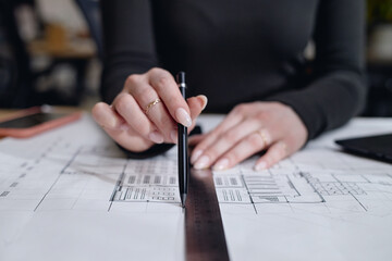 Caucasian young adult woman drawing architectural plans using ruler and pen on desk, focusing on precision while working on technical drawings, partial body visible, hands in foreground