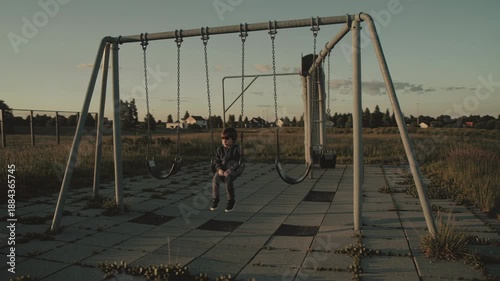 Boy on a swing set moving from stationary to gently swinging in an empty park at sunset. Childhood melancholia and loneliness concept.