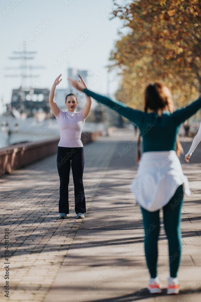Obraz premium A woman stands facing the camera with arms raised while another person participates in a front-facing stretch along a riverside walkway.