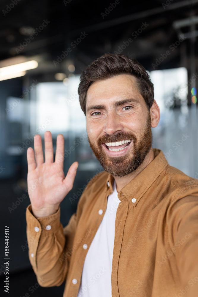 Fototapeta premium Happy man smiling and waving at camera during a friendly video call in a modern office, close-up headshot conveying connection, positivity, remote work and professional casual style