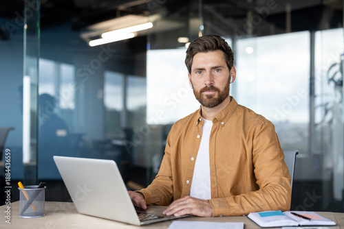 Wallpaper Mural Man with beard and wireless earbuds engaging with his laptop, sitting at a desk in a contemporary office setting, representing focused work and innovative technology use Torontodigital.ca