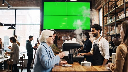 A barista serves coffee to a smiling elderly woman in a busy modern cafe with a green video screen on transparent background