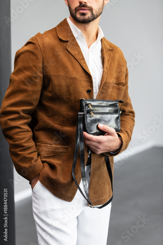 Stylish man in a brown suede blazer and white trousers leaning against a wall holding a small dark leather crossbody bag