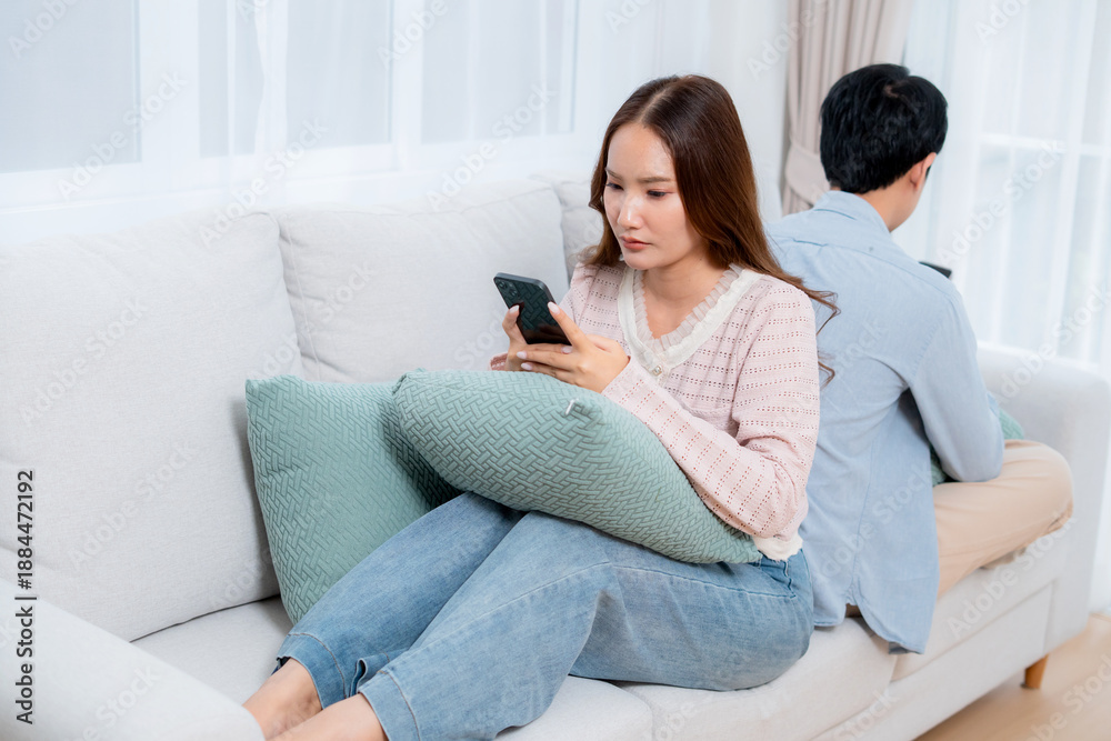 Obraz premium Unhappy asian couple having relationship problem, woman looking upset while man ignoring partner for smartphone, conflict and silence on sofa at home, social media addiction, stress and tension.