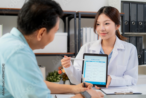 Asian nutritionist using tablet to explaining personalized meal plan with AI to patient during healthcare consultation, healthy lifestyle, woman dietitian showing nutrition plan with AI on tablet.