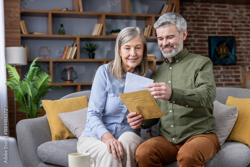 Fotografi Older couple smiling while sitting on a couch in their living room, opening a le