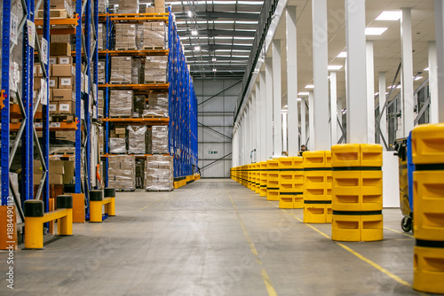Wide warehouse aisle with tall storage racks stacked with pallets of various cardboard boxes, showcasing organised industrial logistics and inventory management.