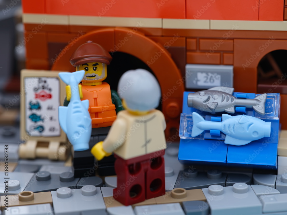 Obraz premium Tambov, Russian Federation - January 19, 2026 Lego fisherman minifigure selling a light blue fish from a fish stall to a customer on a city street.