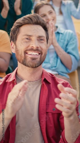 Caucasian handsome young male laughing while celebrating football moment in stadium crowd. Cheerful man in red shirt smiling wide surrounded by clapping fans. Excited reaction during game.