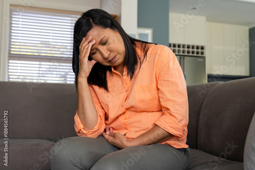 Middle-aged Asian woman sitting on gray couch at home in orange shirt holding head and stomach