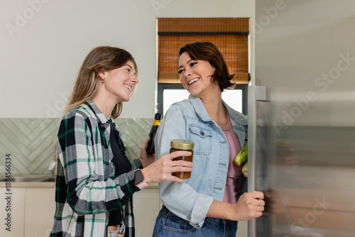 Diverse female friends chatting by steel fridge in kitchen, holding vegetables, beer, tumbler