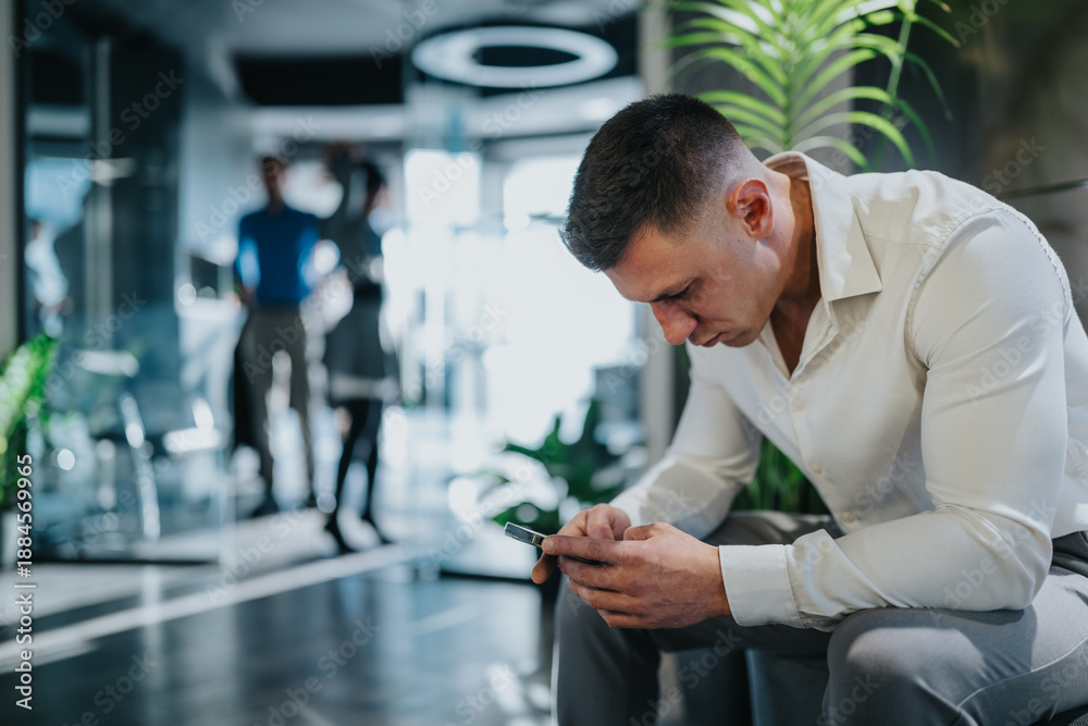 Fototapeta premium A focused man in a white shirt uses a smartphone in a modern office lobby. The scene shows him checking messages while blurred colleagues chat in the background.