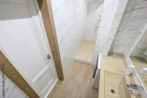 Foto A modern, compact bathroom with marble-patterned tiles, wooden flooring, white door, built-in shower, beige countertop, and sleek fixtures