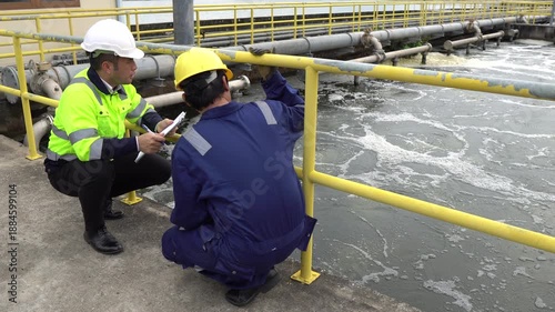Two Asian engineers discussing maintenance at wastewater treatment plant. They check water quality and pipeline system outdoors. Industrial ecology and environmental management concept.