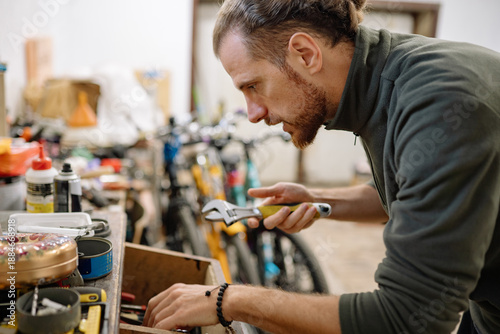 Male mechanic with long hair selecting the right wrench from cluttered toolbox in home garage workshop.