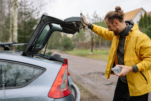 Man opens car trunk on a cloudy day while holding a water bottle near a dirt road surrounded by trees