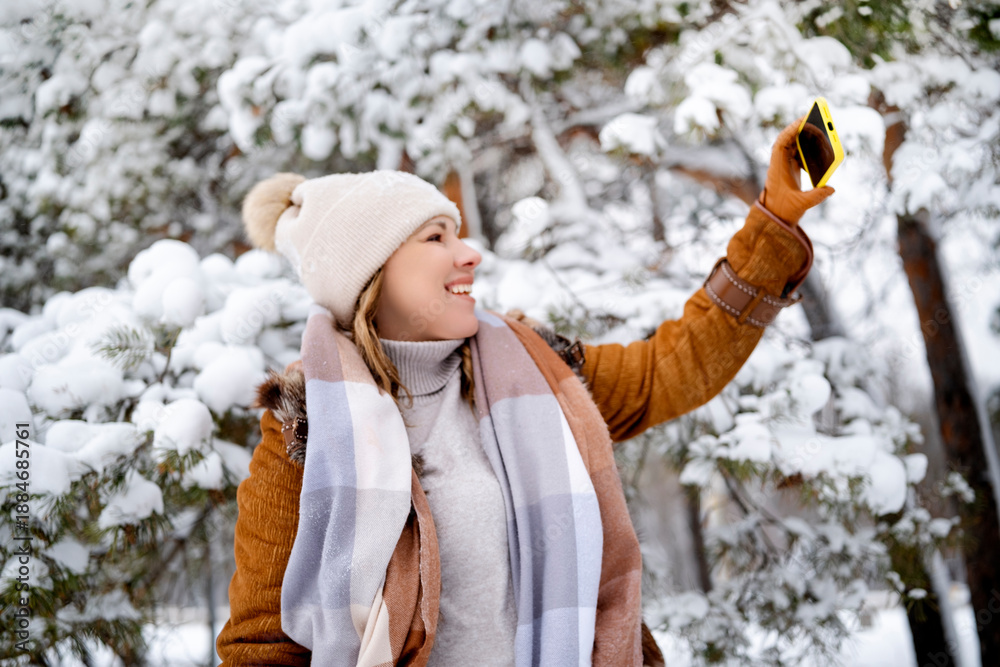 Fototapeta premium Joyful adult female takes selfie with smartphone in snowy pine forest. Woman in brown coat near trees with snow. Blogging, technology, winter, travel, lifestyle concept