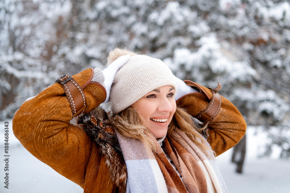 Fototapeta premium Radiant woman laughs and holds head in snowy winter park. Middle aged female in brown sheepskin jacket and white hat. Joy, happiness, weekend, nature, lifestyle concept