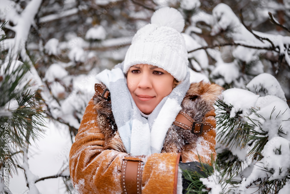 Fototapeta premium Thoughtful woman in warm coat and white hat looks away in snowy forest. Winter mood portrait. Middle aged female near trees with snow. Nature, relax, lifestyle concept