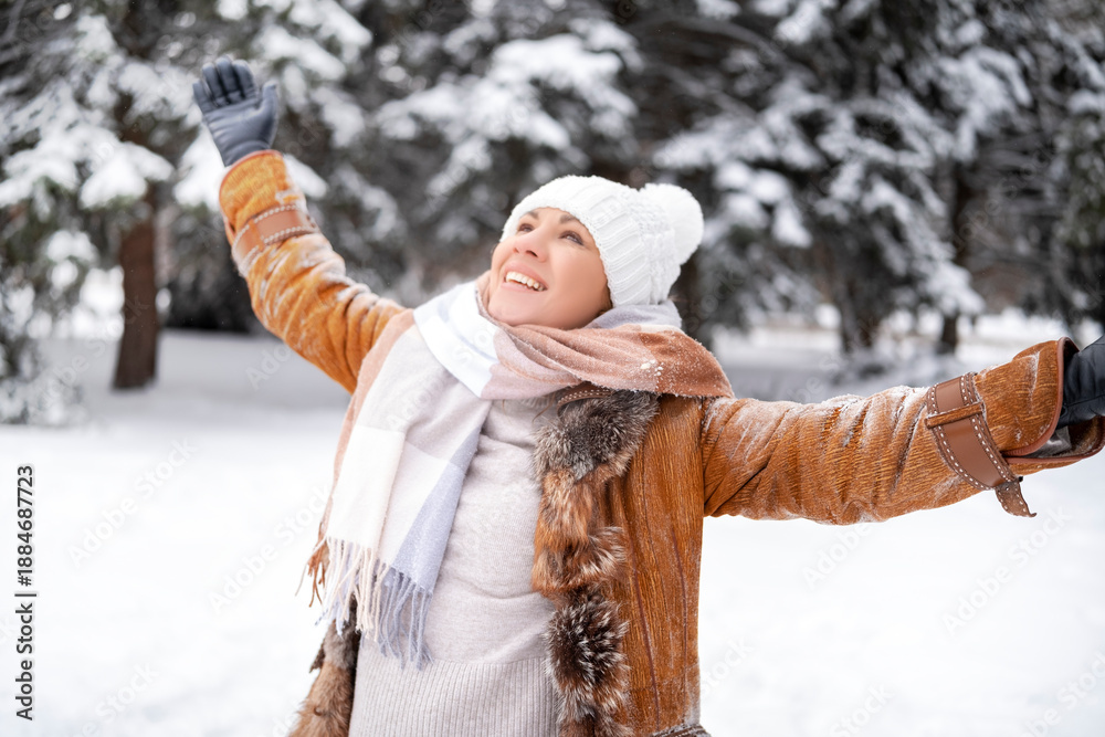 Fototapeta premium Joyful woman raises hands and looks up in snowy winter forest. Happiness and freedom. Middle aged female in brown coat and white hat. Nature, wellness, lifestyle concept