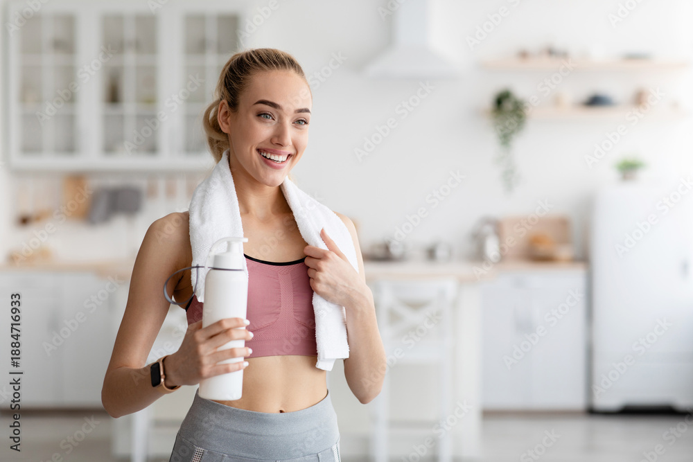 Obraz premium Covid-19 lockdown, body and health care, thirst and coach recommendations. Cheerful young pretty blonde lady in sportswear with towel on shoulders holds bottle of water in minimalist kitchen interior