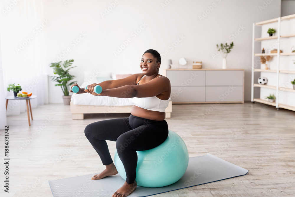 Fototapeta premium Plus size black woman working out with dumbbells on fitness ball at home, empty space. Curvy African American lady losing weight, having domestic training, leading healthy lifesyle, indoors