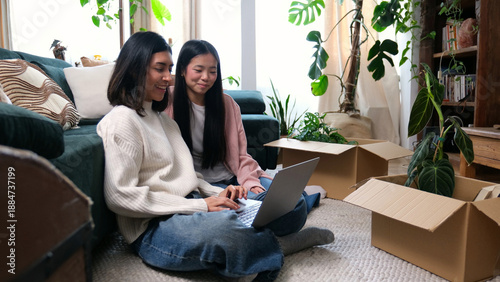 Tableau sur toile Happy women using a laptop while unpacking plants and boxes in their new apartme