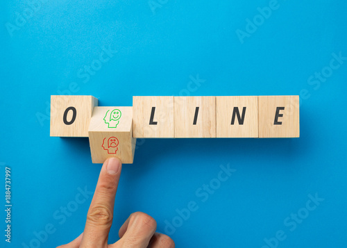 A finger flips a wooden block to choose between a happy or sad face icon within the word ONLINE on a blue background. Concept for mental health, social media impact, and digital wellbeing balance.