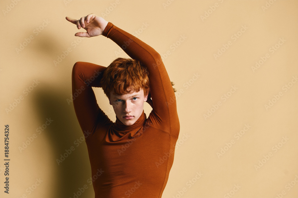 Obraz premium Portrait of young redhead woman posing with arms raised wearing brown turtleneck sweater on beige background with serious expression and direct gaze.