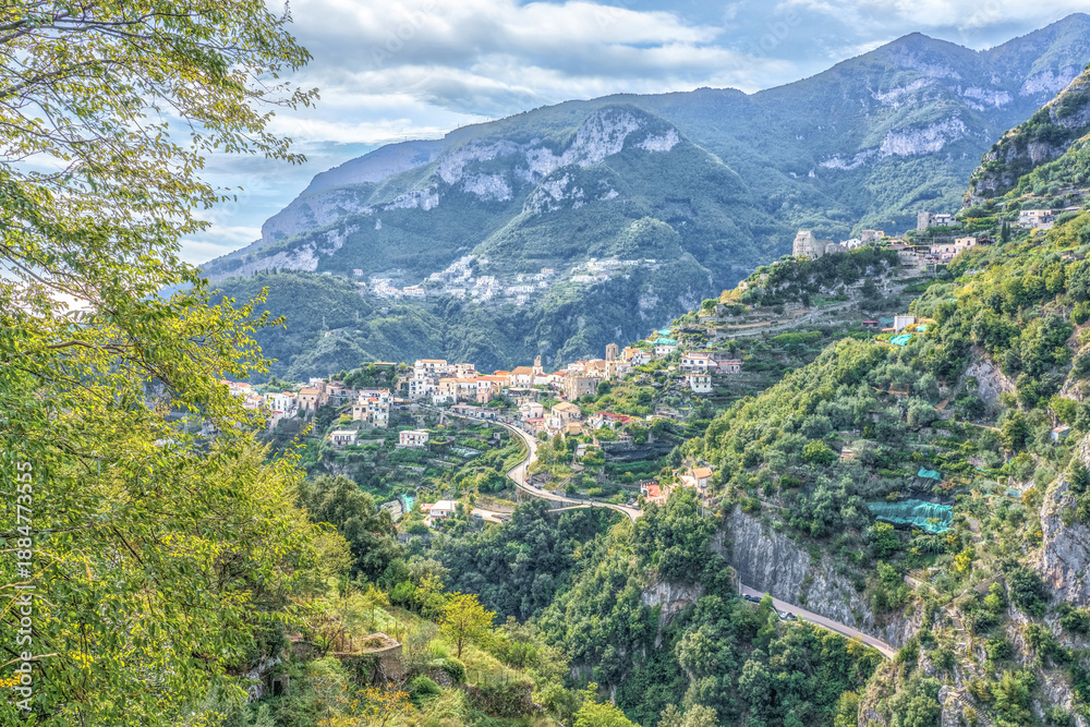 Fototapeta premium Amalfi Coast, Italy. Scenic view of a hillside village nestled among lush green mountains with winding roads and a cloudy sky in the background during daylight hours