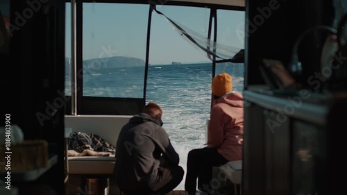 Man and woman sit on stern of yacht watching turbulent wake left by motor. View of bay surrounded by steep cliffs. Back view. Slow motion