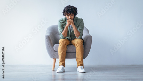 Fényképezés A man with curly hair is sitting on a gray chair in a room with plain walls